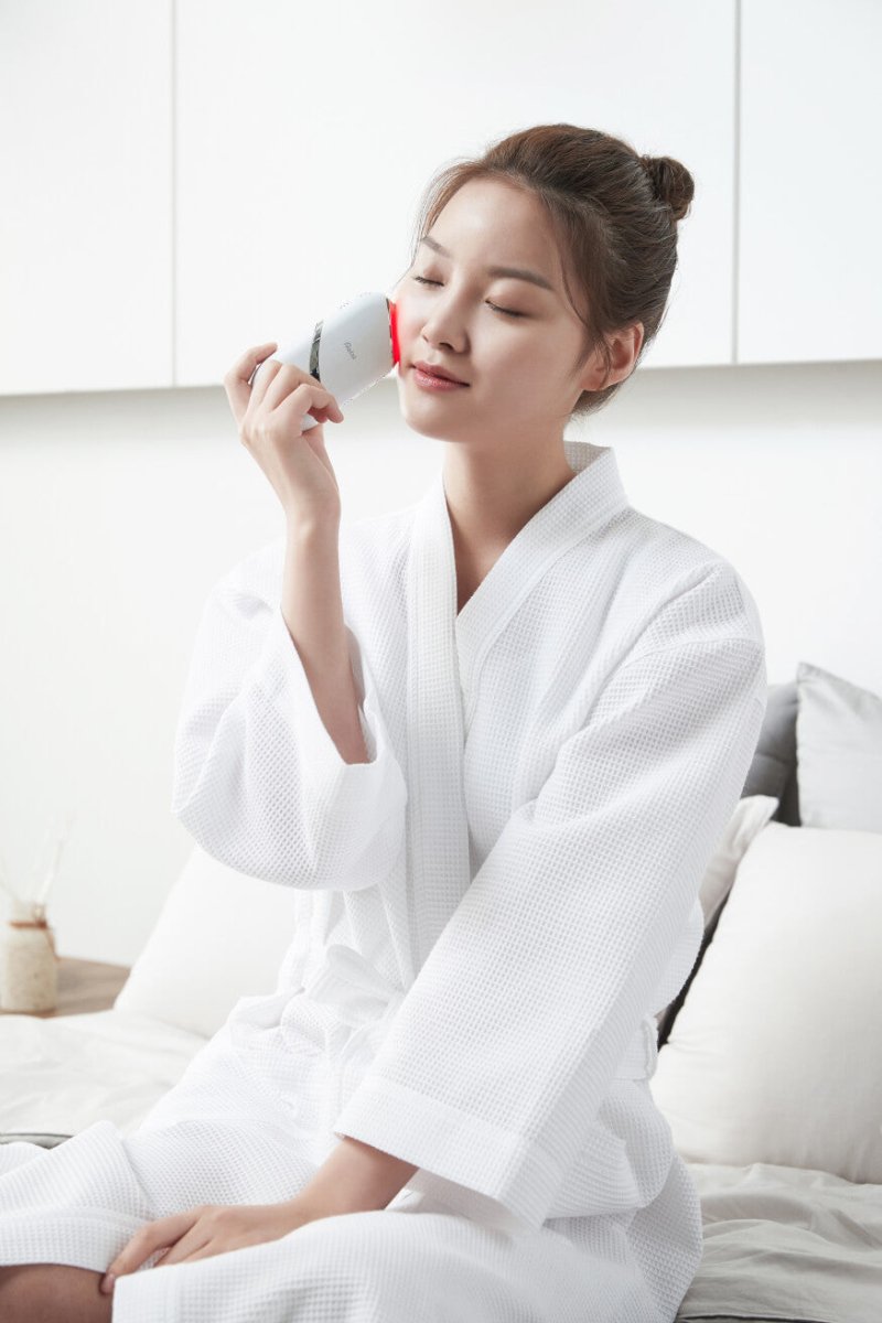A woman in a white robe using Aalok Snowfit on her face in a bright room.